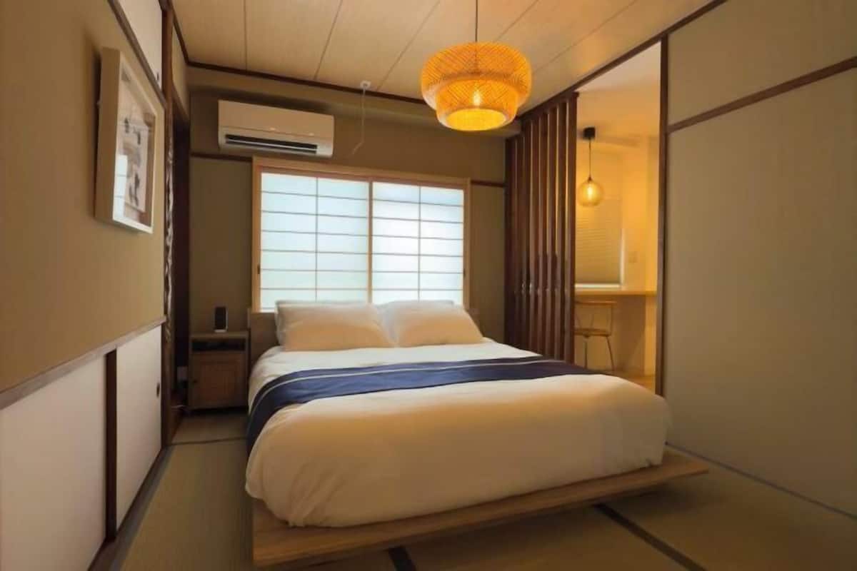 A traditional Japanese-style bedroom with tatami flooring, a bed with a dark blue bedspread, a hanging lamp, and a window with shoji screens.