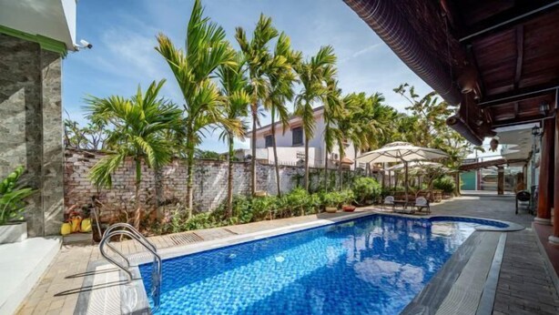 Une piscine bordée d’un mur en pierre et entourée de palmiers, avec une terrasse couverte.