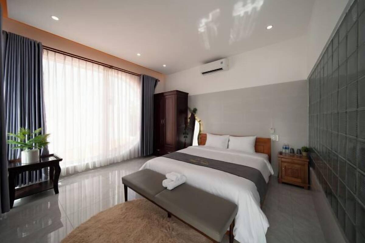 A hotel room with a large bed, a bench, a bedside table, a wardrobe, and a large window with curtains.