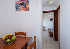 A dining area with a table, chairs, a fruit bowl, and a hallway leading to a bedroom with a bed and a painting.