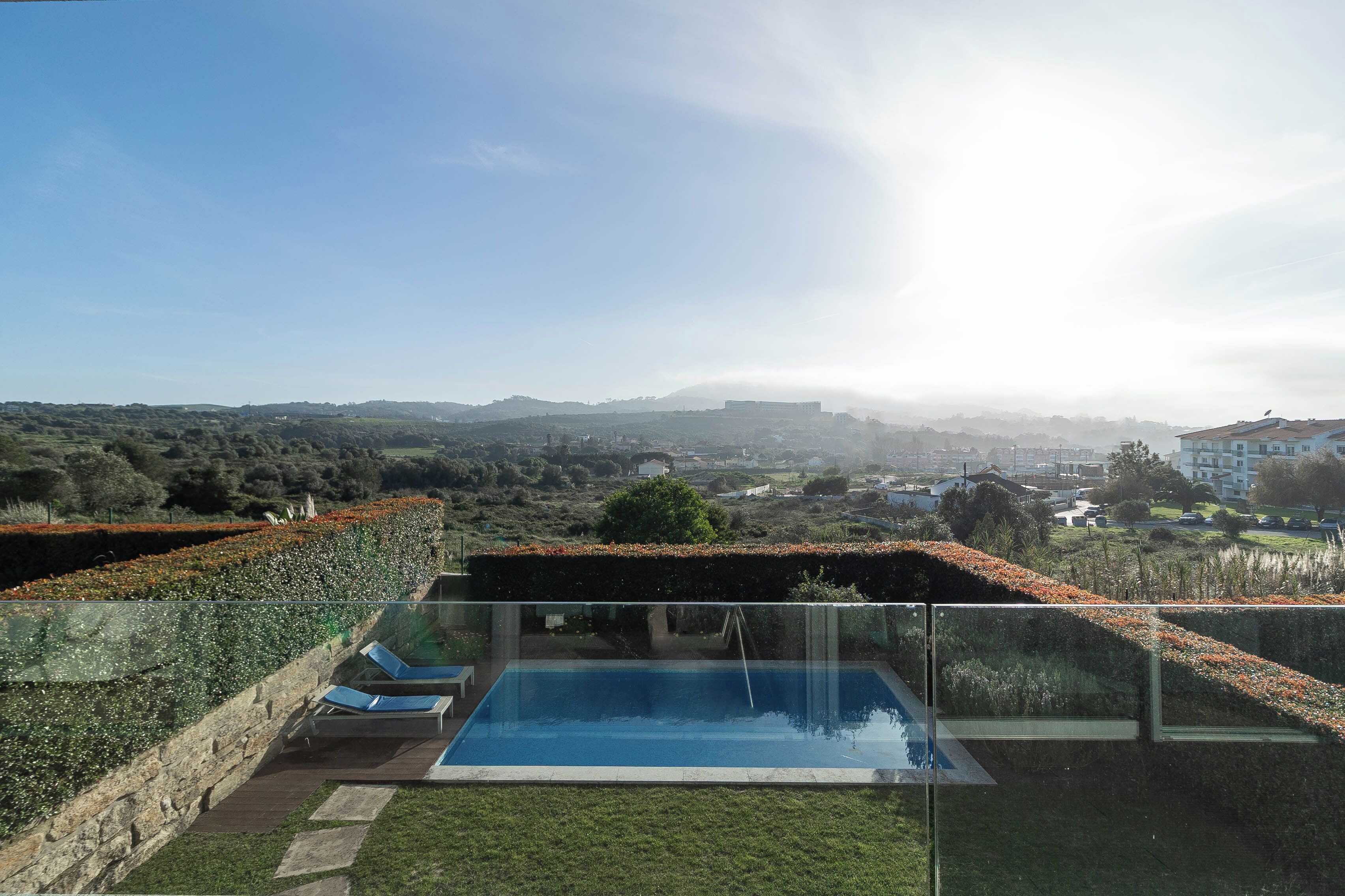 Une piscine extérieure moderne, dotée de parois en verre transparent, entourée d’un jardin bien entretenu et offrant une vue sur une ville lointaine.