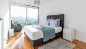 A modern bedroom with a large bed, a bedside table, a chair, and a view of the city.