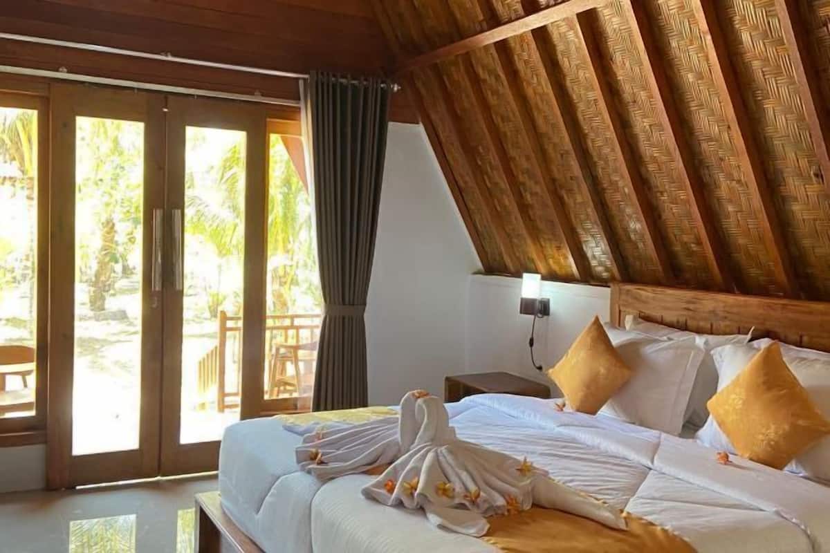 A hotel room with a bed, pillows, a nightstand, and a view through a sliding door.