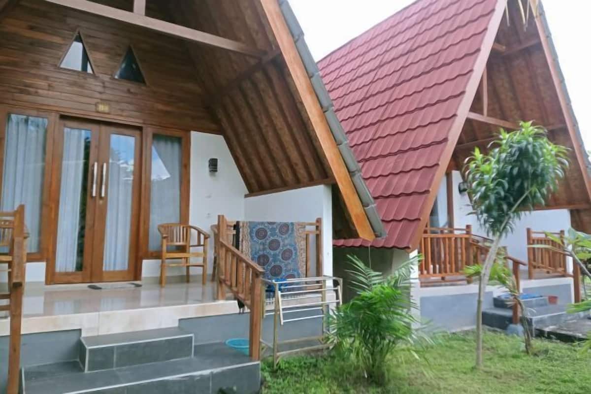 A wooden house with a red roof, a porch with chairs, and a small garden.