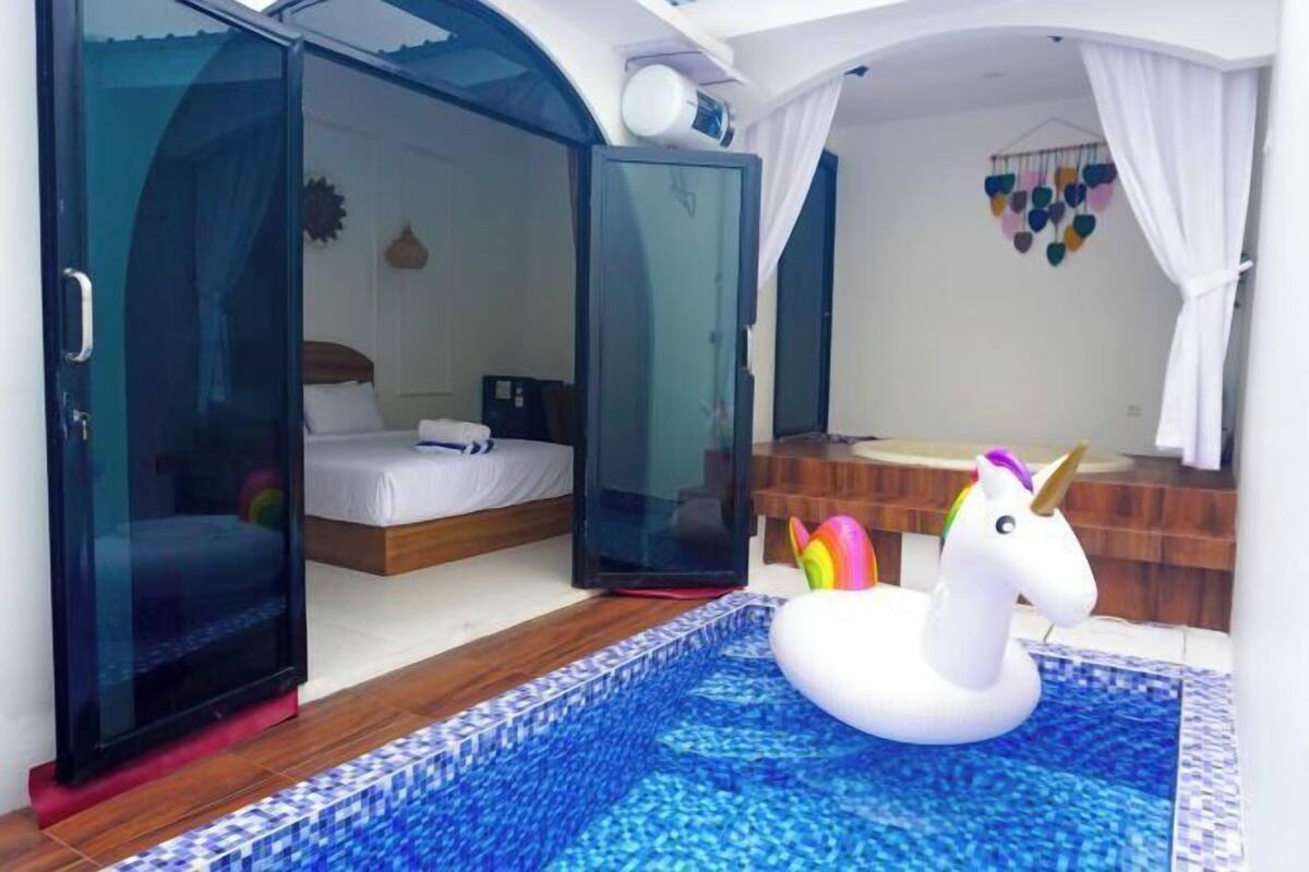 Een slaapkamer met een glazen deur, een bed, een zwembadje met een eenhoorn opblaasbaar speeltje, en een badkamer met een spiegel en een hangende decoratie.