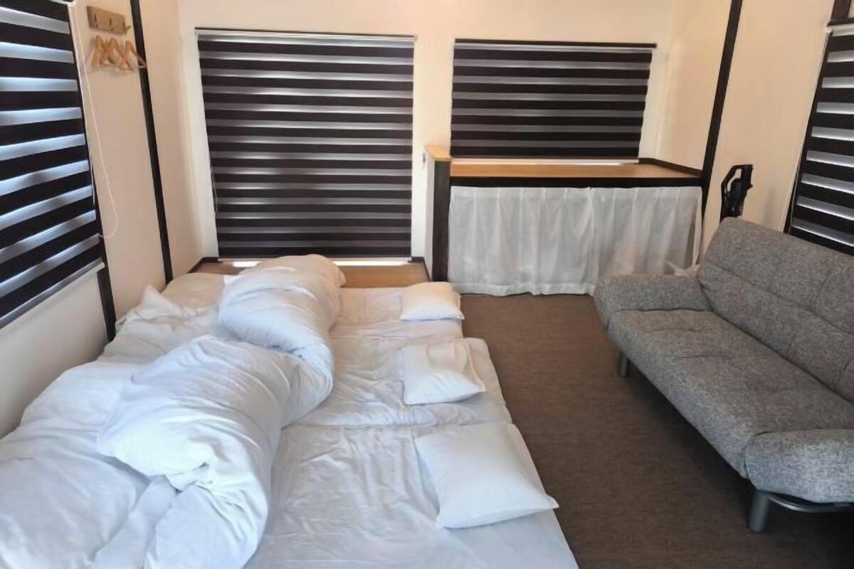 A compact room with a bed, a grey sofa, and two window blinds.