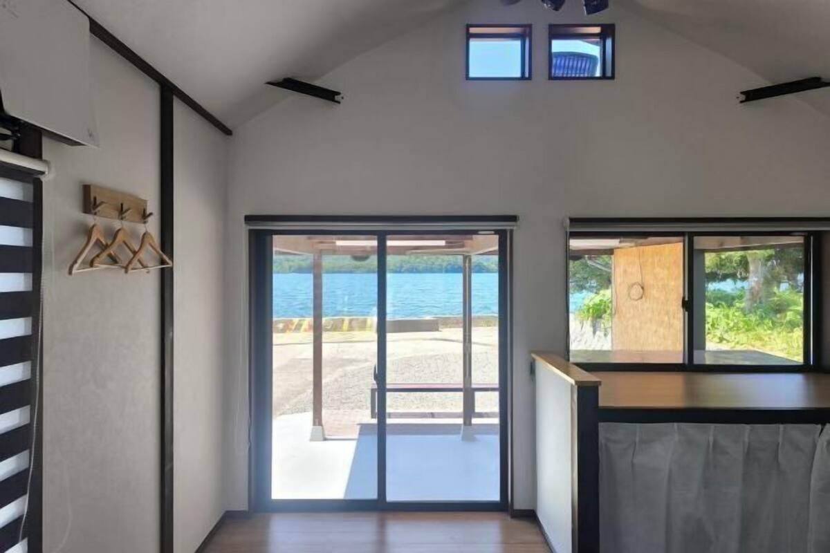 A room with a large sliding glass door leading to a beach, a small window with a view, and a wooden bench.