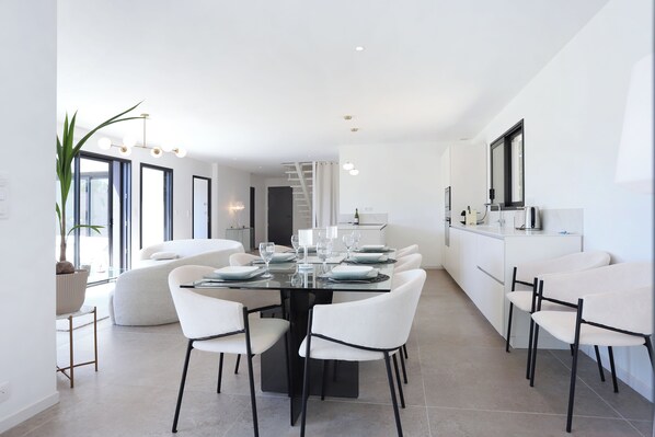 Uma área de refeições moderna, com mesa de vidro, cadeiras brancas e uma cozinha ao fundo.