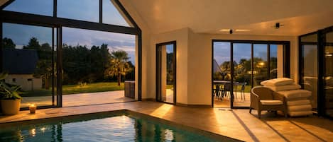 Une piscine intérieure moderne, dotée d’une eau claire, de grandes fenêtres et d’un espace patio aménagé avec du mobilier d’extérieur.