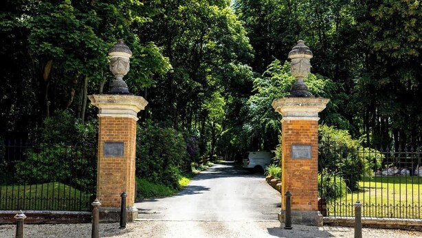 Un camino pavimentado que conduce a una entrada porticada con dos pilares de ladrillo coronados por jarrones decorativos.