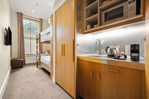 A compact hotel room with a bunk bed, kitchenette, and a large window with blinds.