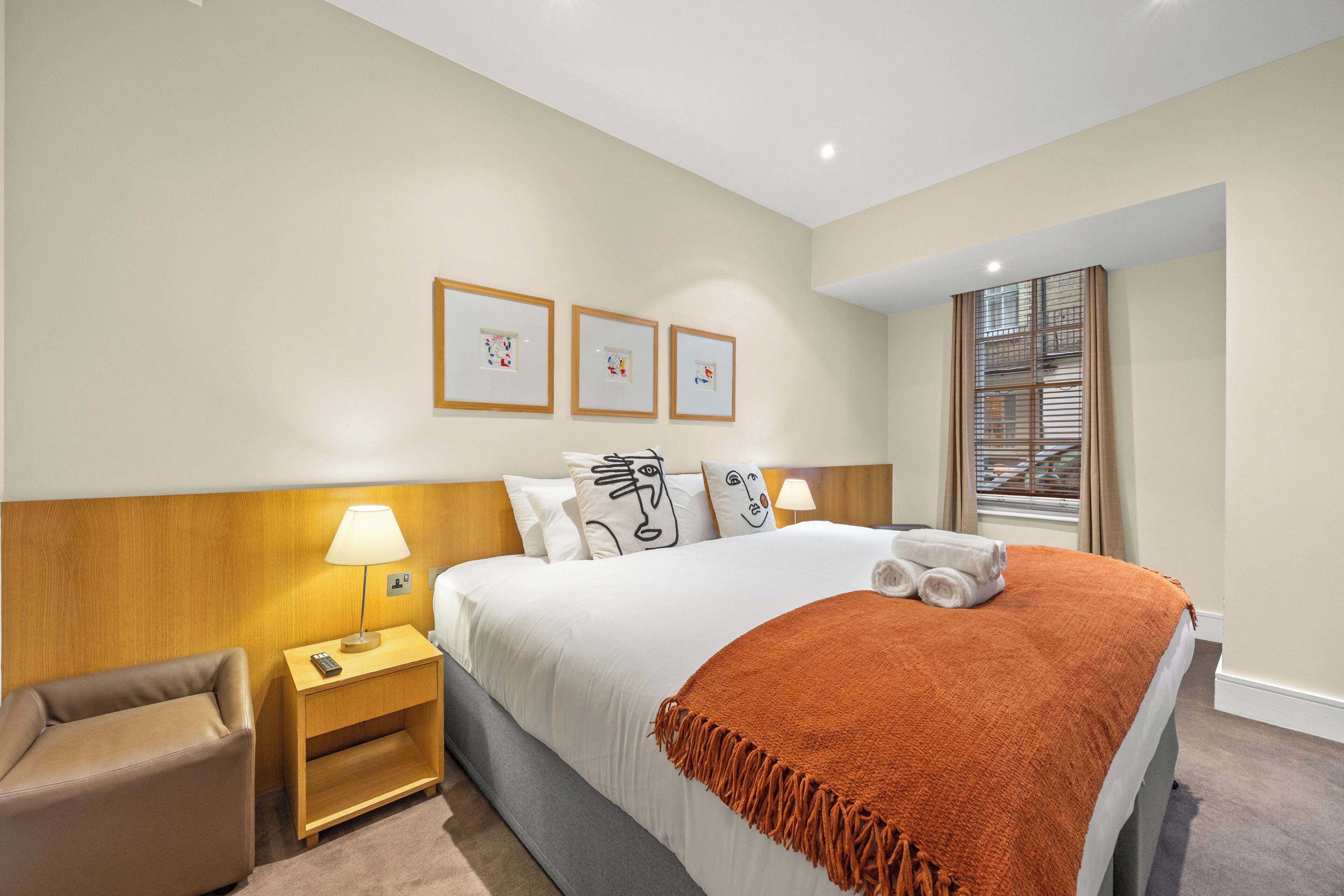 A hotel room with a bed, a nightstand, a chair, a window with blinds, and wall-mounted artwork.