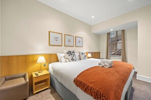 A hotel room with a bed, a nightstand, a chair, a window with blinds, and wall-mounted artwork.