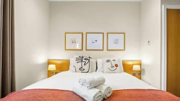 A hotel room with a bed, a red blanket, white towels, three framed artworks, two bedside lamps, and a wooden headboard.