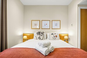 Ein Hotelzimmer mit einem Bett, einer roten Decke, weißen Handtüchern, drei gerahmten Kunstwerken, zwei Nachttischlampen und einem hölzernen Kopfteil.