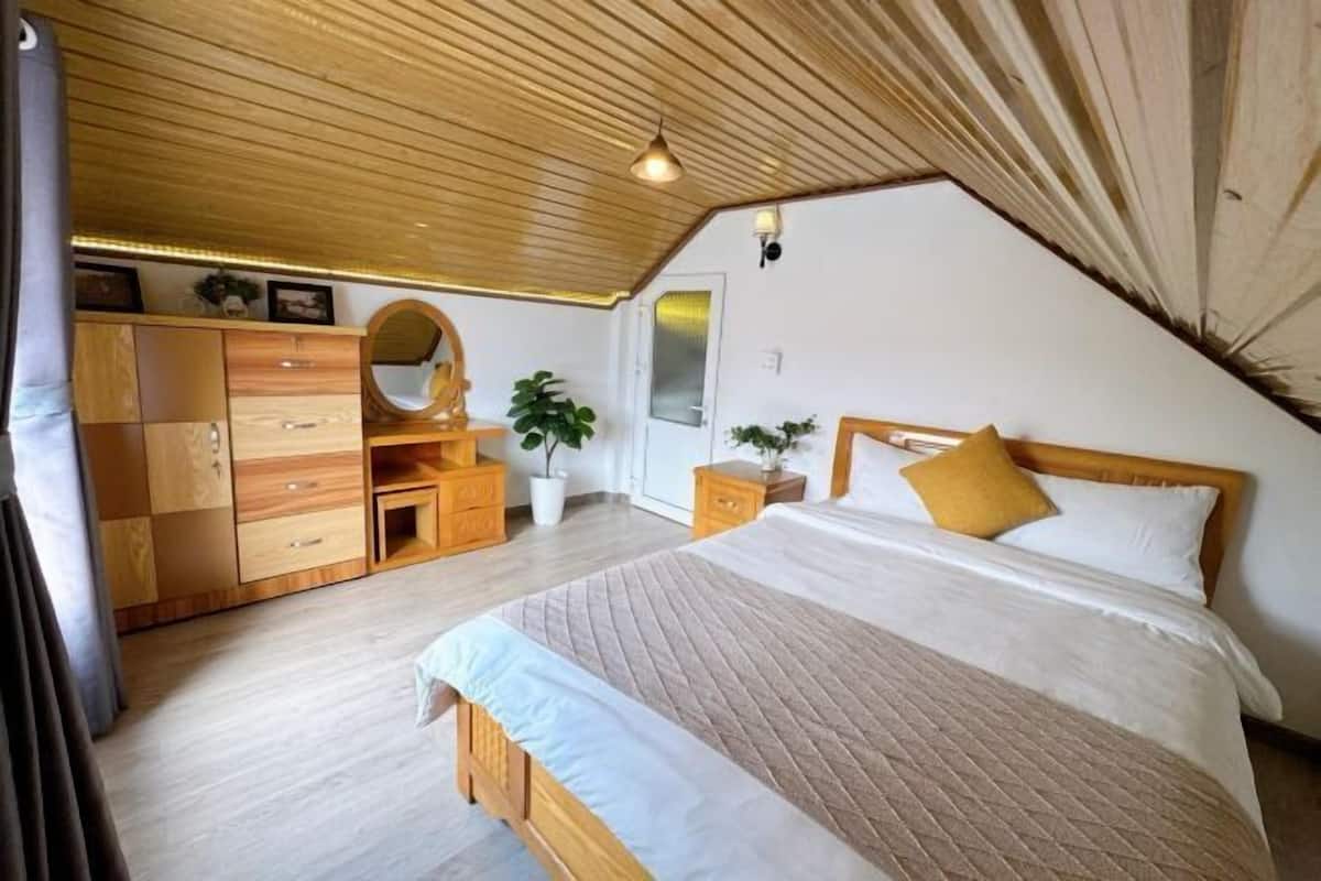 A bedroom with a wooden ceiling, a bed with white and beige bedding, a wooden dresser, a mirror, and a potted plant.