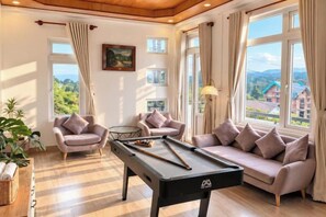 A pool table in a room with a sofa and armchairs, large windows with curtains, and a painting on the wall. - Tuong Vy Villa (Dalat)