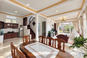 A spacious dining area with a wooden table and chairs, a staircase leading to an upper level, and a view of the outdoors through large windows. - Tuong Vy Villa (Dalat)