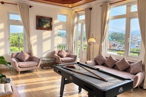 A pool table in a room with two sofas and a table, large windows with curtains, and a painting on the wall. - Tuong Vy Villa (Dalat)