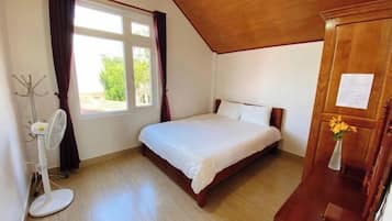 A bedroom with a bed, a window with curtains, a standing fan, and a wooden dresser.