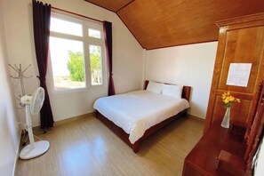 A bedroom with a bed, a window with curtains, a standing fan, and a wooden dresser. - Tuong Vy Villa (Dalat)