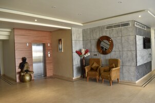 Um hall de entrada com uma mesa de recepção, duas poltronas, uma estátua de Buda, um espelho fixado na parede e um elevador moderno.