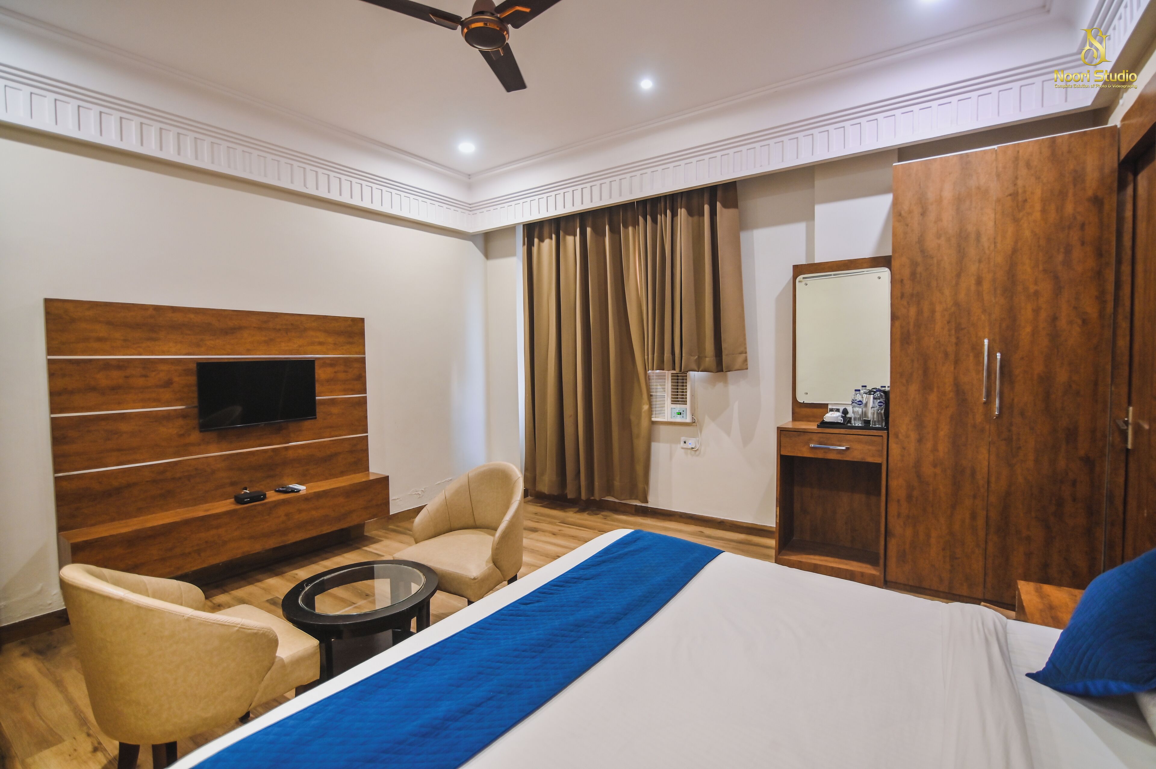 A hotel room with a bed, a TV mounted on a wooden panel wall, a desk with a chair, a wardrobe, and a ceiling fan.