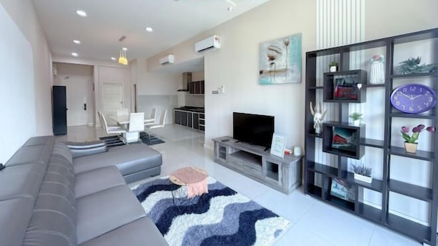 A modern living room with a grey sectional sofa, a flat-screen TV, and a decorative shelf with plants and a clock. - The Loft Imago Shopping Mall By Chill Time (Kota Kinabalu)