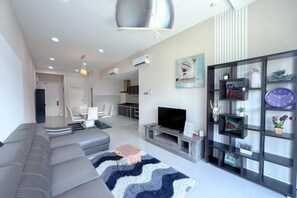 Een moderne woonkamer met een grijze hoekbank, een flatscreen-televisie en een decoratieve plank met planten en een klok.