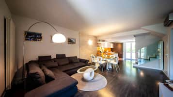 Un salon moderne avec un canapé d’angle marron foncé, une table basse ronde et un coin repas comprenant une table avec un plateau en verre et des chaises blanches.