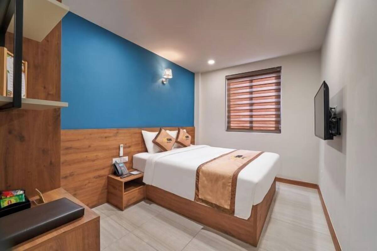 Habitación de hotel con cama, mesita de noche, escritorio, televisor montado en la pared y ventana con persianas.