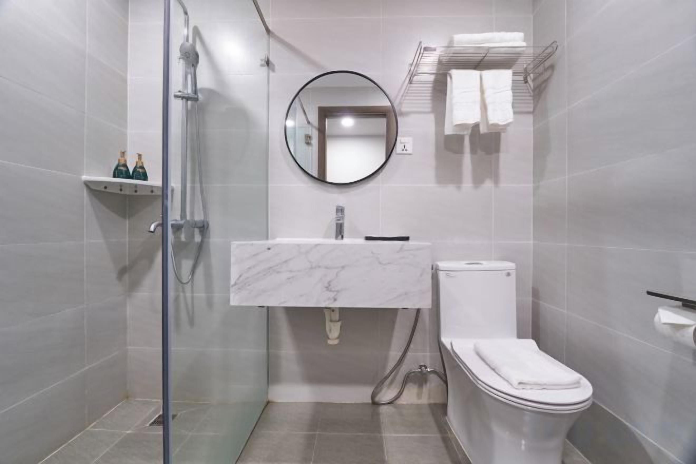 A modern bathroom with a glass shower enclosure, a white marble sink, a toilet, and a round mirror.