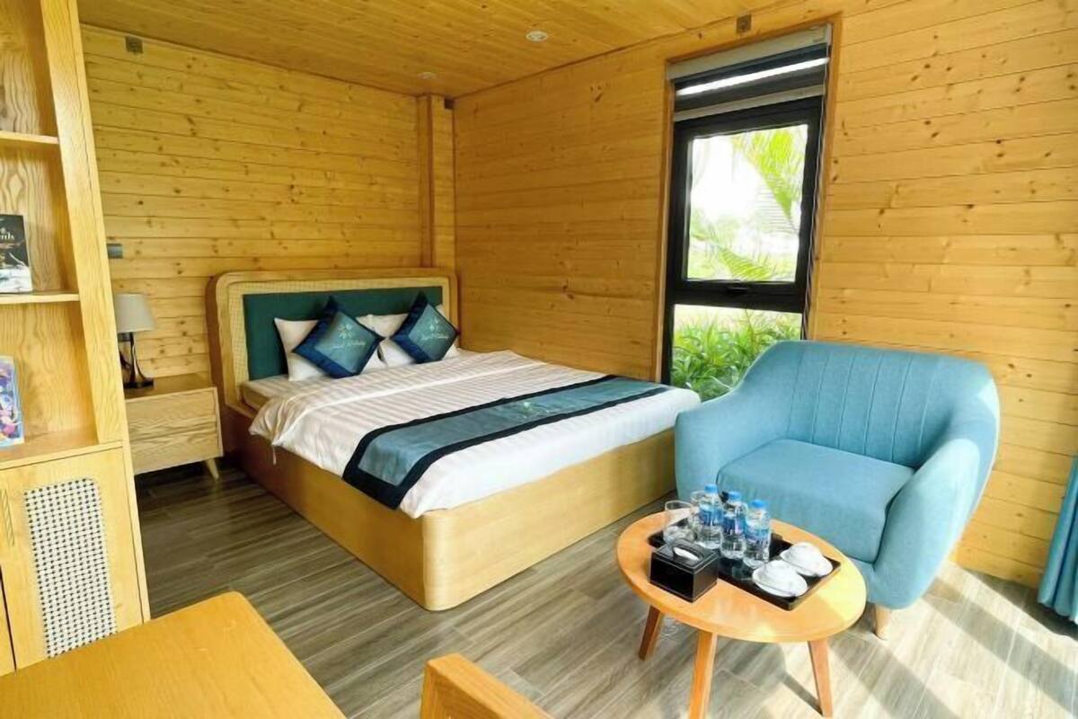 Una habitación de madera con una cama, un sillón azul, una mesita con detalles y una ventana con follaje en el exterior.