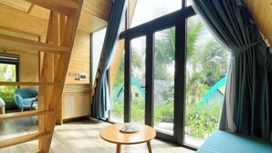 Una habitación con piso de madera, una mesa redonda con un cuenco, ventanas amplias con cortinas y vista a la vegetación.