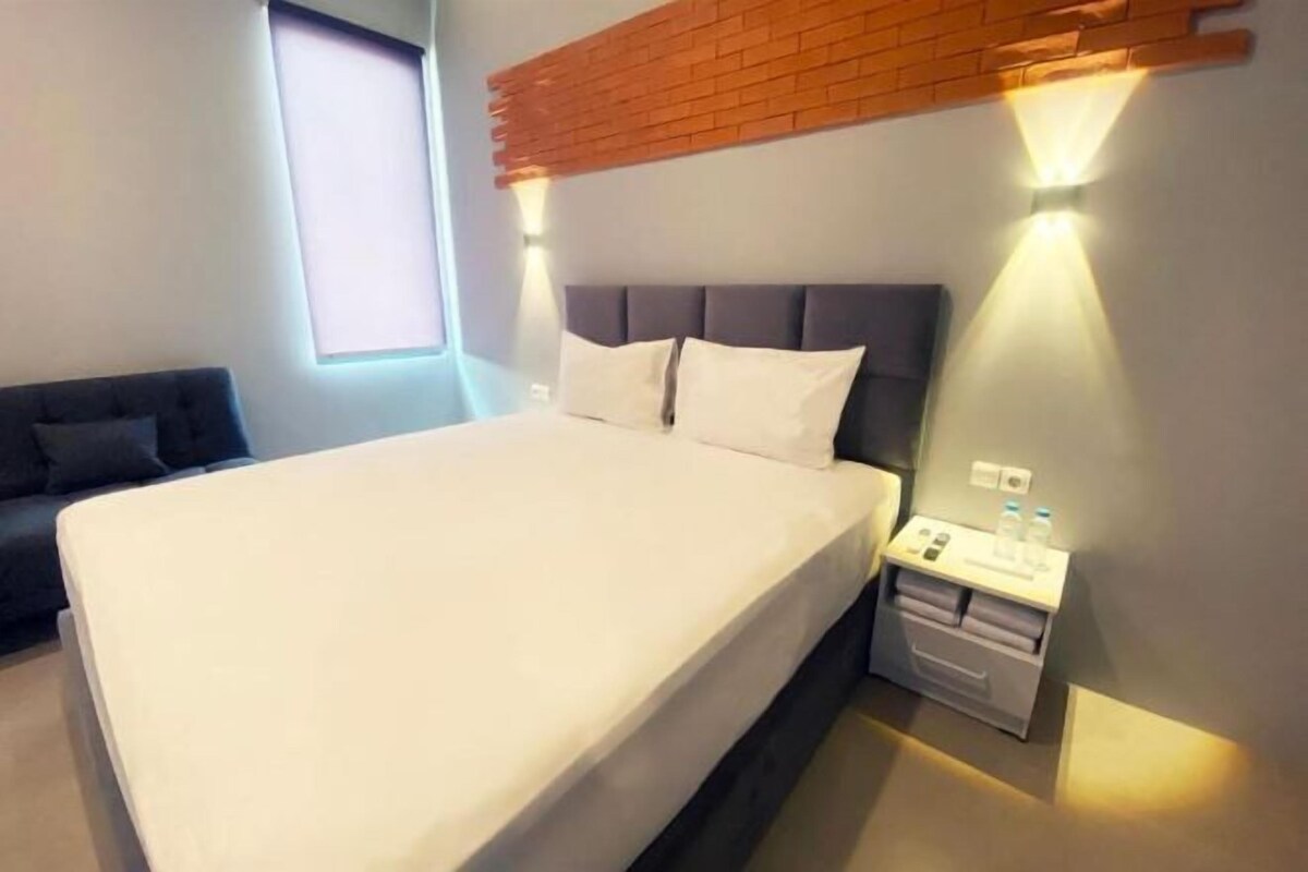 A modern hotel room with a large bed, a bedside table with a lamp, a small nightstand, and a window with blinds.