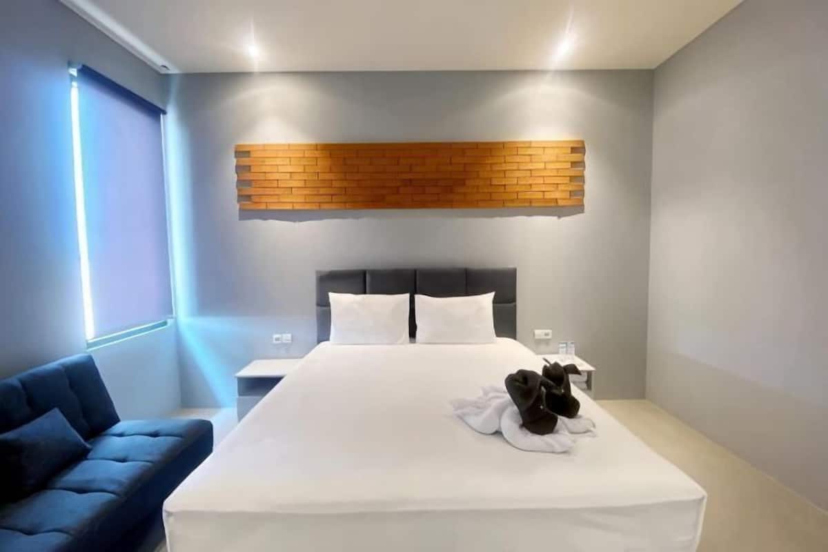 Quarto de hotel moderno com uma cama grande, um sofá azul, uma parede de tijolos e uma mesa de cabeceira com luminária.
