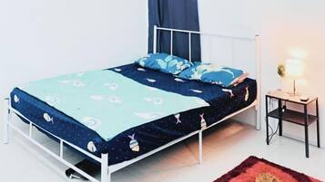 Una cama individual con estructura metálica blanca, colcha de diseño azul y blanco y almohadas a juego. Hay una mesita de noche pequeña con una lámpara y una alfombra roja en el suelo.