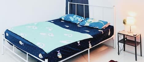 Habitación con una cama individual de estructura metálica blanca, colcha con estampado azul y blanco, cortina azul oscuro, mesita de noche con lámpara y alfombra roja.
