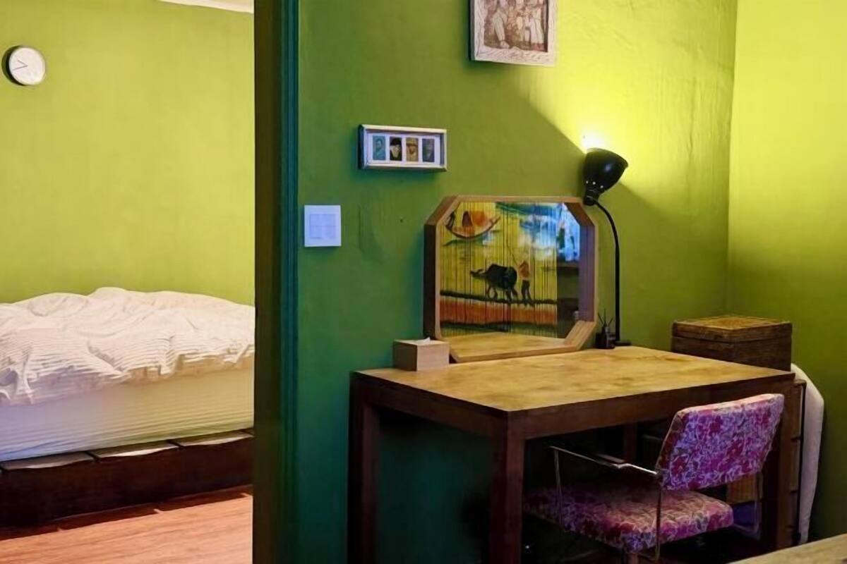 Een kamer met een bed, een houten tafel met een stoel, een lamp en een lijst met een afbeelding aan de muur.