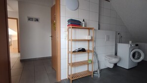 A bathroom with a toilet, washing machine, and a wooden shelf with folded clothes and a bottle. - Apartment 'Ferienwohnung Haak' with Private Terrace, Balcony and Wi-Fi (Dörpen)
