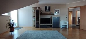 Ein Wohnzimmer mit Fernseher, Regalen, einer Topfpflanze und einem weißen Sessel.