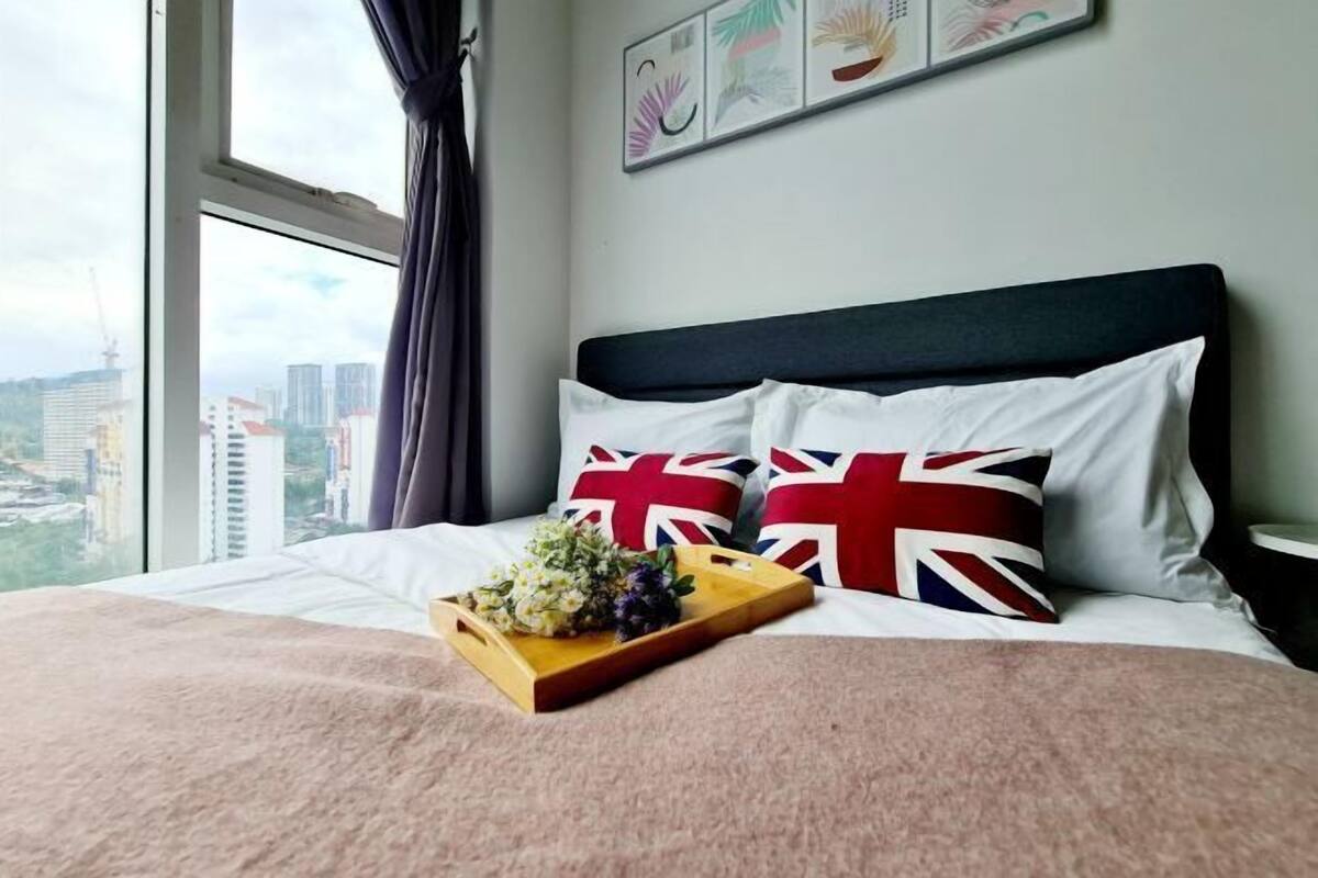 A bed with a wooden tray of flowers, a view of buildings through a window, and a wall with framed artwork.