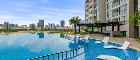 A swimming pool with lounge chairs, a city skyline in the background, and a high-rise building.