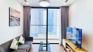 A modern living room with a sofa, a TV on a wooden stand, a painting on the wall, and a large window with curtains. - Vinhomes Skylake by TA Home – Premium Hanoi Apartment (Hanoi)