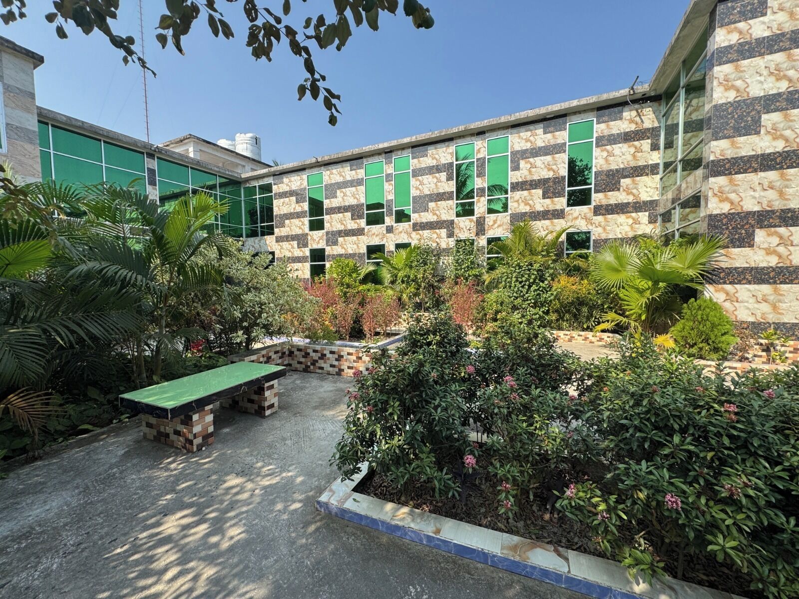 A courtyard with a green bench, surrounded by various plants and a building with patterned stone walls and green windows.