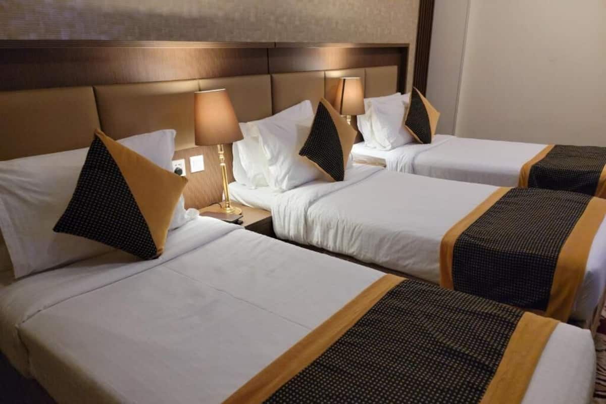 A hotel room with three beds, each with a headboard, white bedding, and a patterned runner. There are bedside lamps on each nightstand.