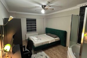 Un dormitorio con cama, escritorio y silla, ventilador de techo, ventana con persianas y zona de ducha.