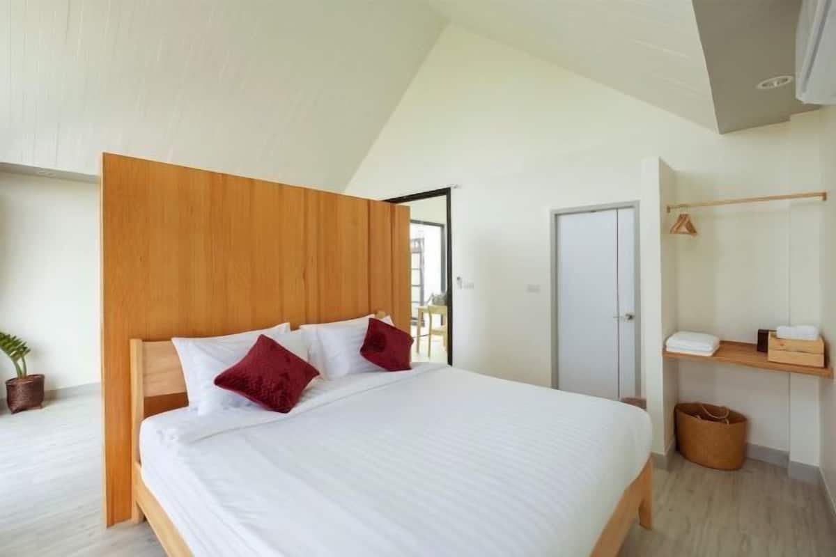 A modern bedroom with a wooden headboard, a large bed with white bedding, a wooden floor, a small plant, and a wooden shelf with towels.