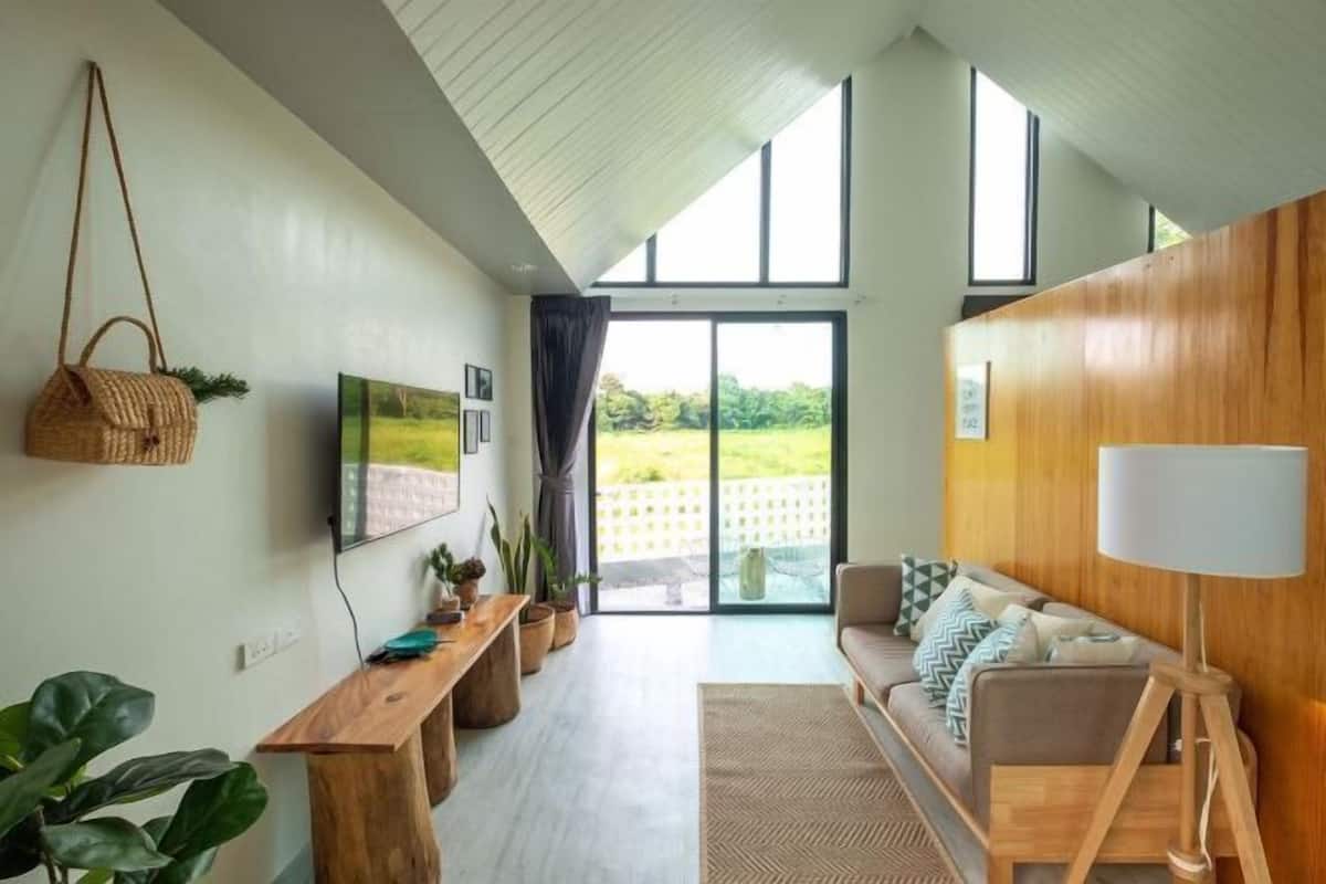 A modern living room with a wooden bench, a sofa, a lamp, and a TV. There is a large window with a view of greenery outside.