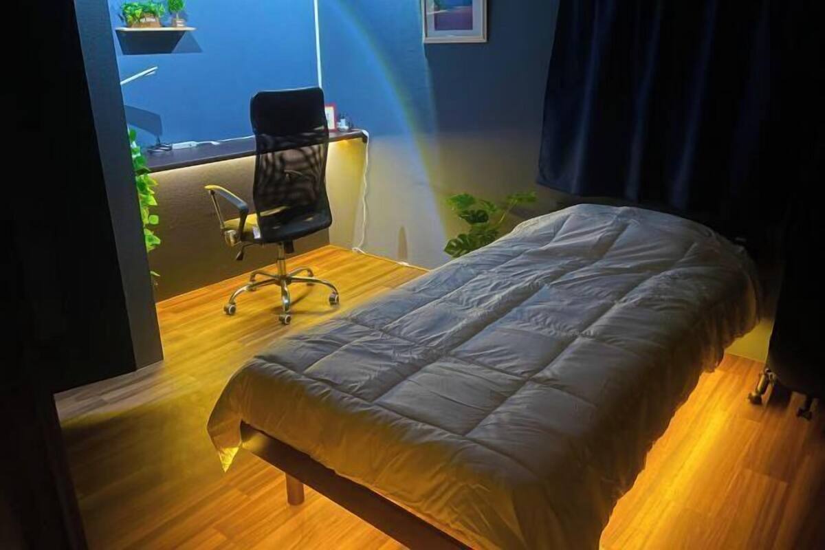 A bedroom with a bed, a desk with a chair, a small plant, and a window with curtains.